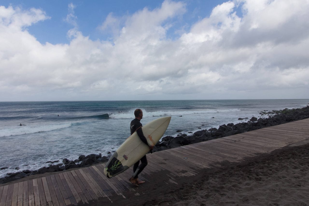 A surfer carrying a board along a seafront walkway as waves break behind