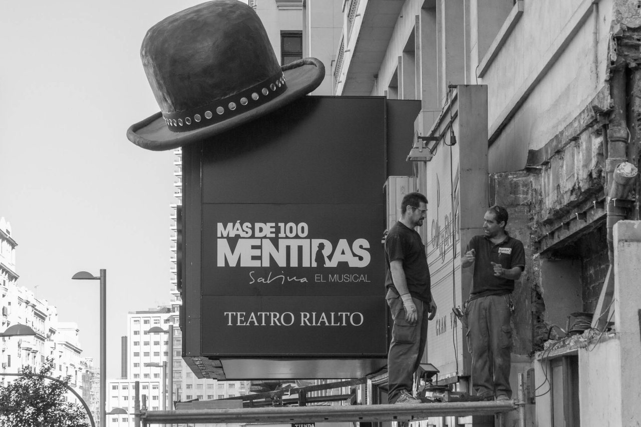 Black and white street scene with a large theatre poster and a bowler hat sign above a passer-by